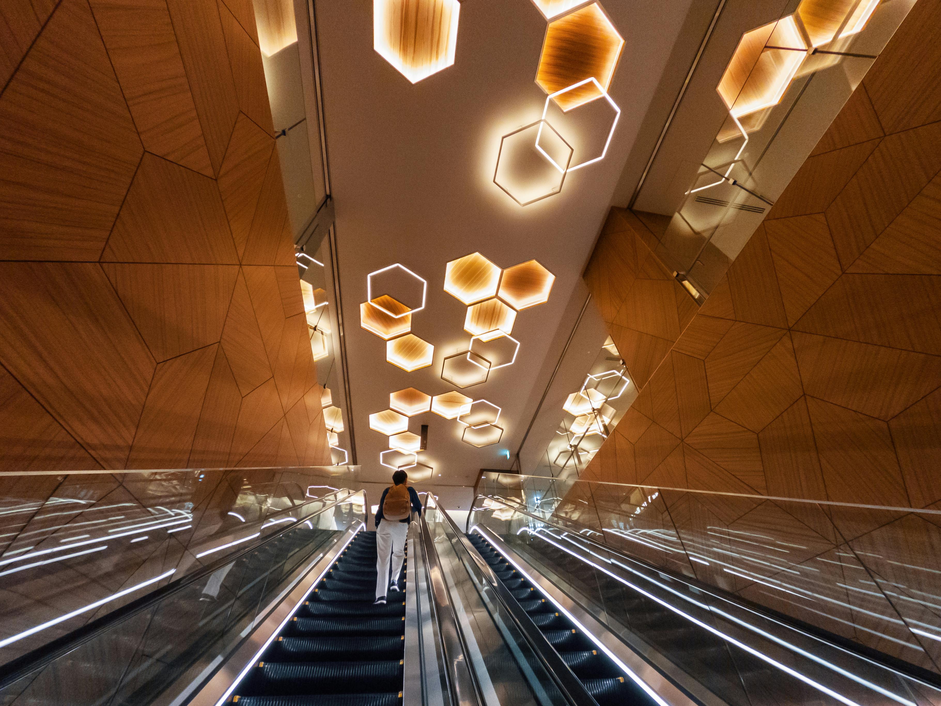 Person ascending an escalator with a modern geometric ceiling design in an urban setting.