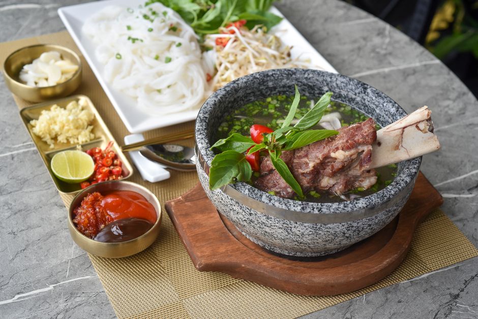 A white bowl of beef pho with rice noodles, thin slices of pink beef, bean sprouts, Thai basil, and lime wedges on the side, steam rising from the clear broth.