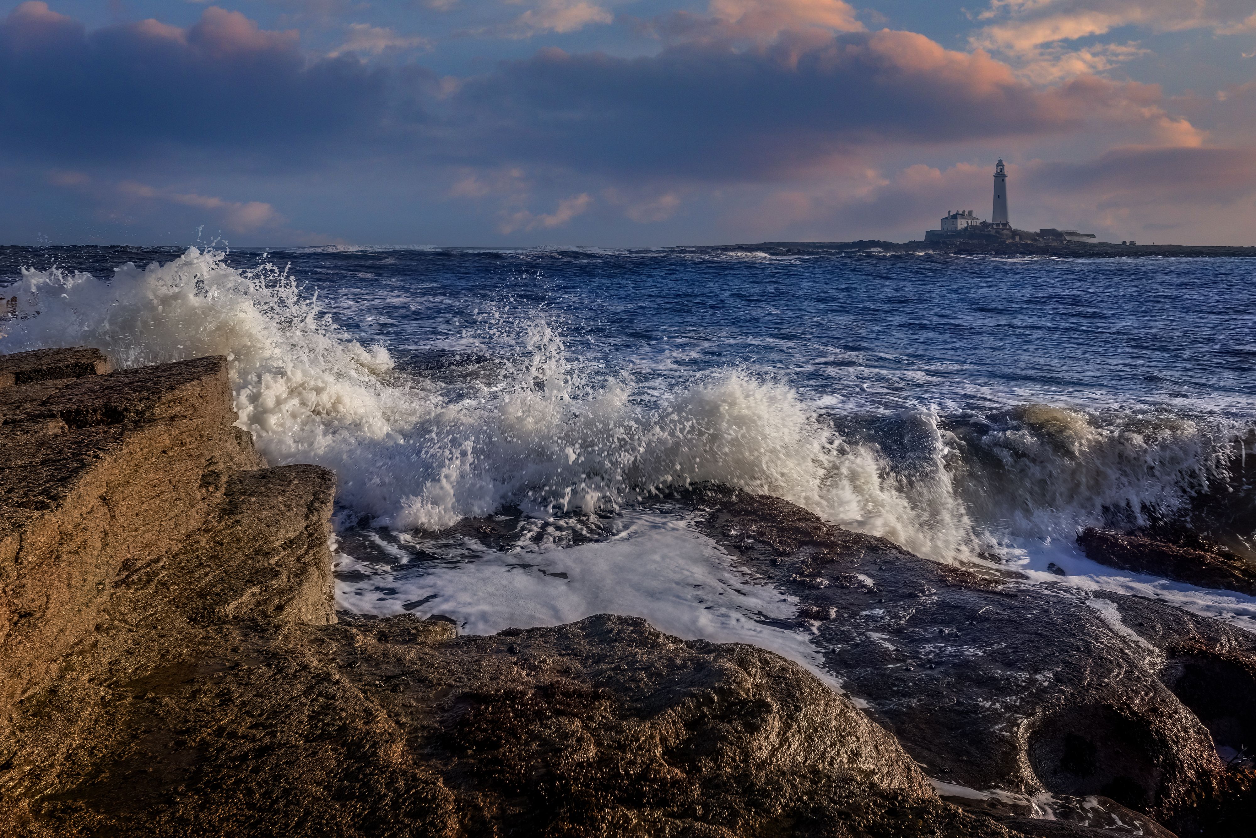 A lighthouse and rocks in the ocean at sunset · Free Stock Photo