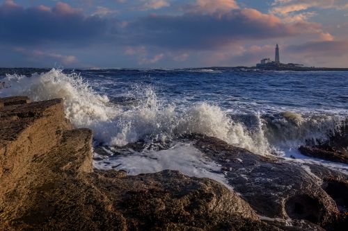 Free Powerful waves crash on rocky coast at sunset with lighthouse in the distance. Stock Photo