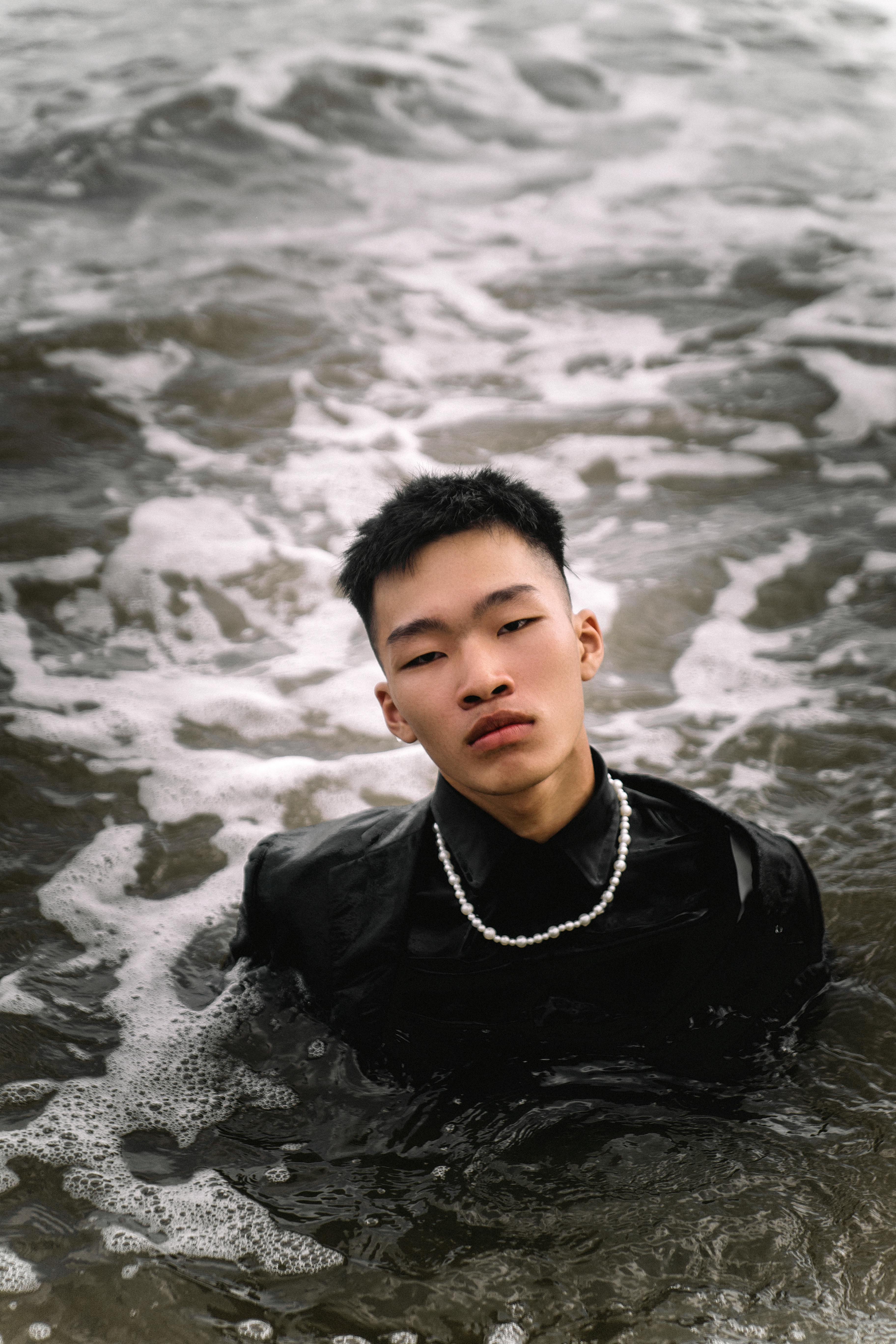 A man stands in waist-deep water wearing a black shirt and pearl necklace, portraying a thoughtful expression.