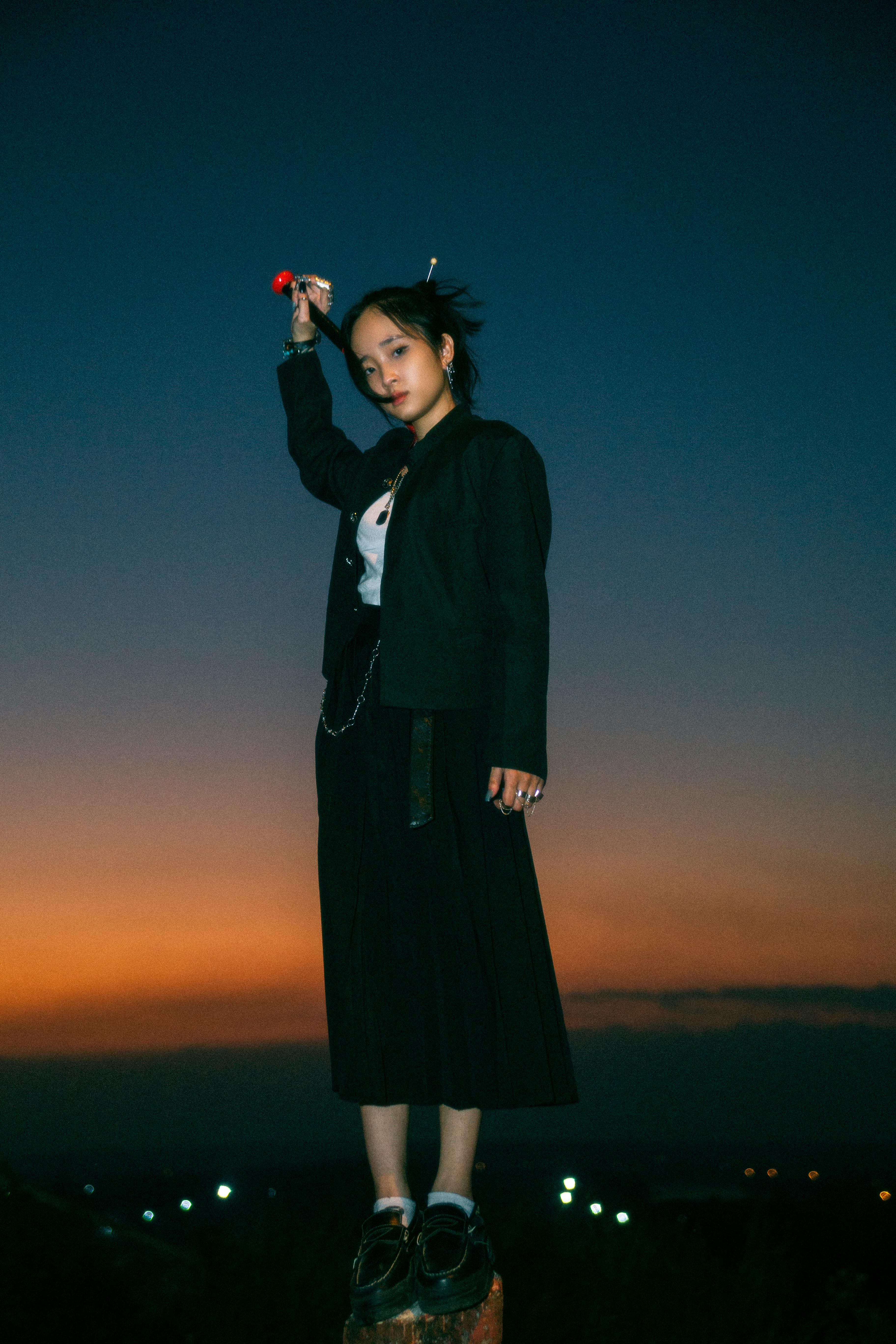 Stylish Asian woman in dark clothing posing at sunset, showcasing modern fashion.