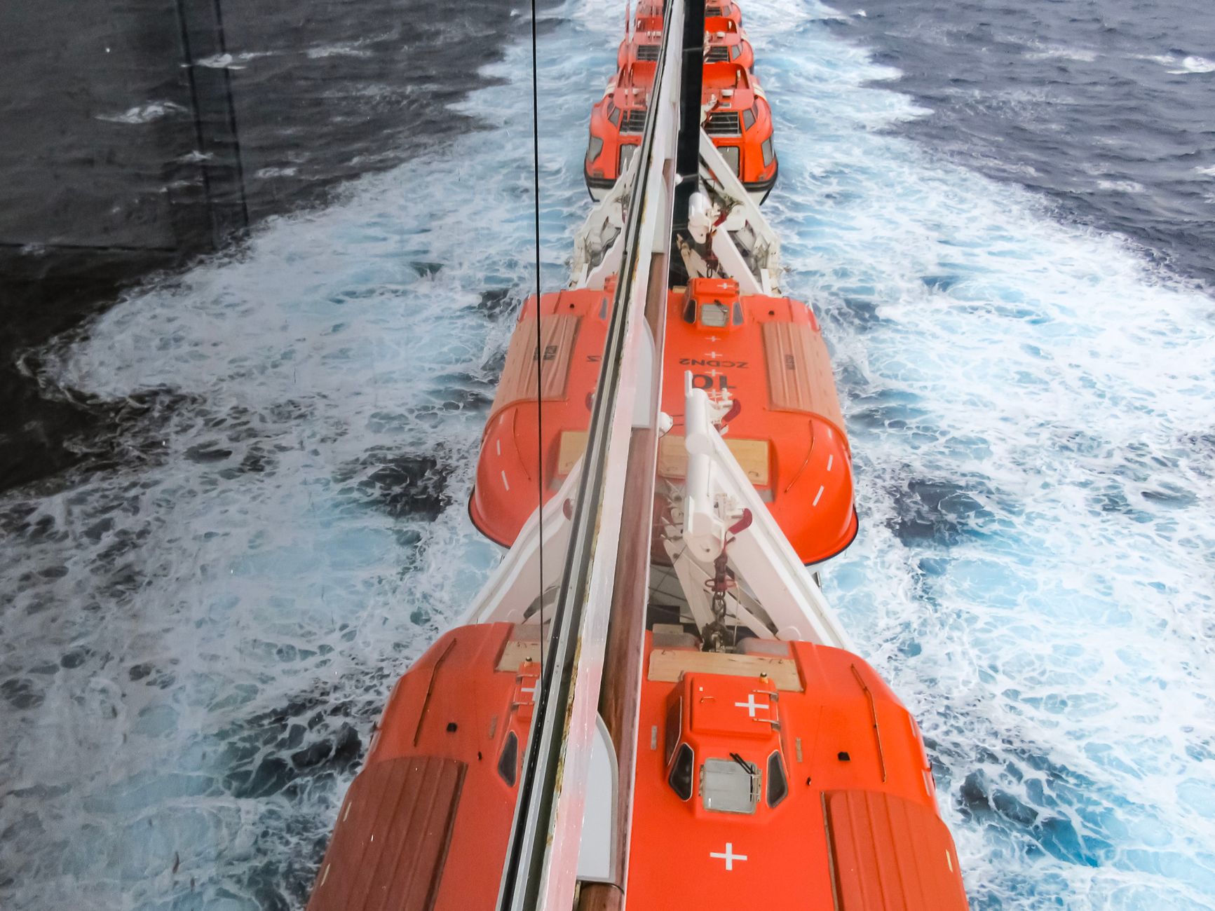 Vibrant orange lifeboats line a cruise ship moving through blue ocean waters.