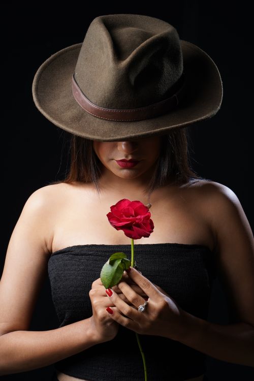 Free A stylish woman in a dark hat holds a red rose against a black backdrop. Stock Photo