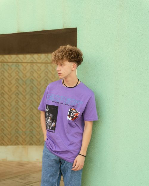Free Fashionable teen boy wearing a purple graphic tee and jeans, leaning against a pastel wall. Stock Photo