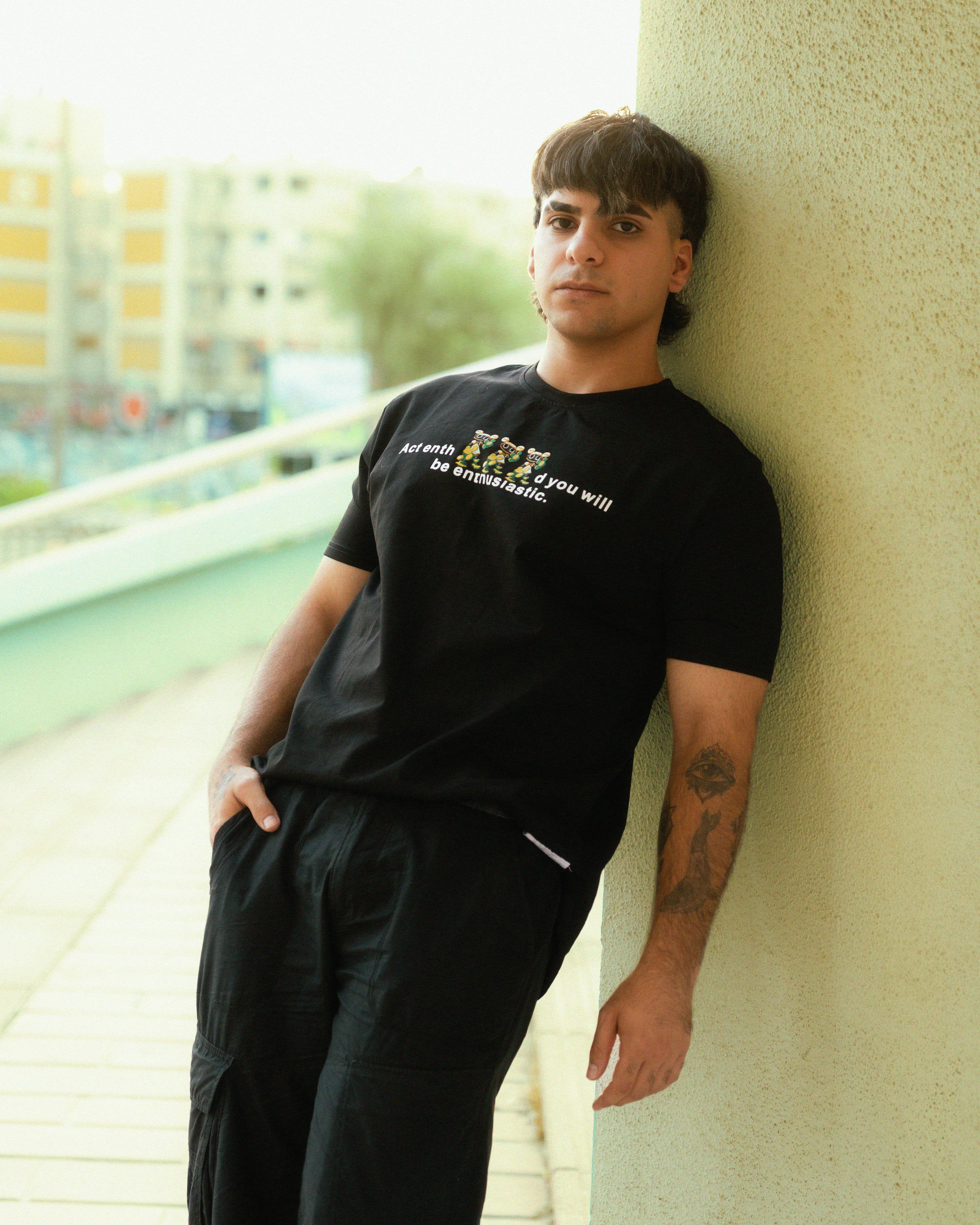 a man leaning against a wall wearing a black t shirt