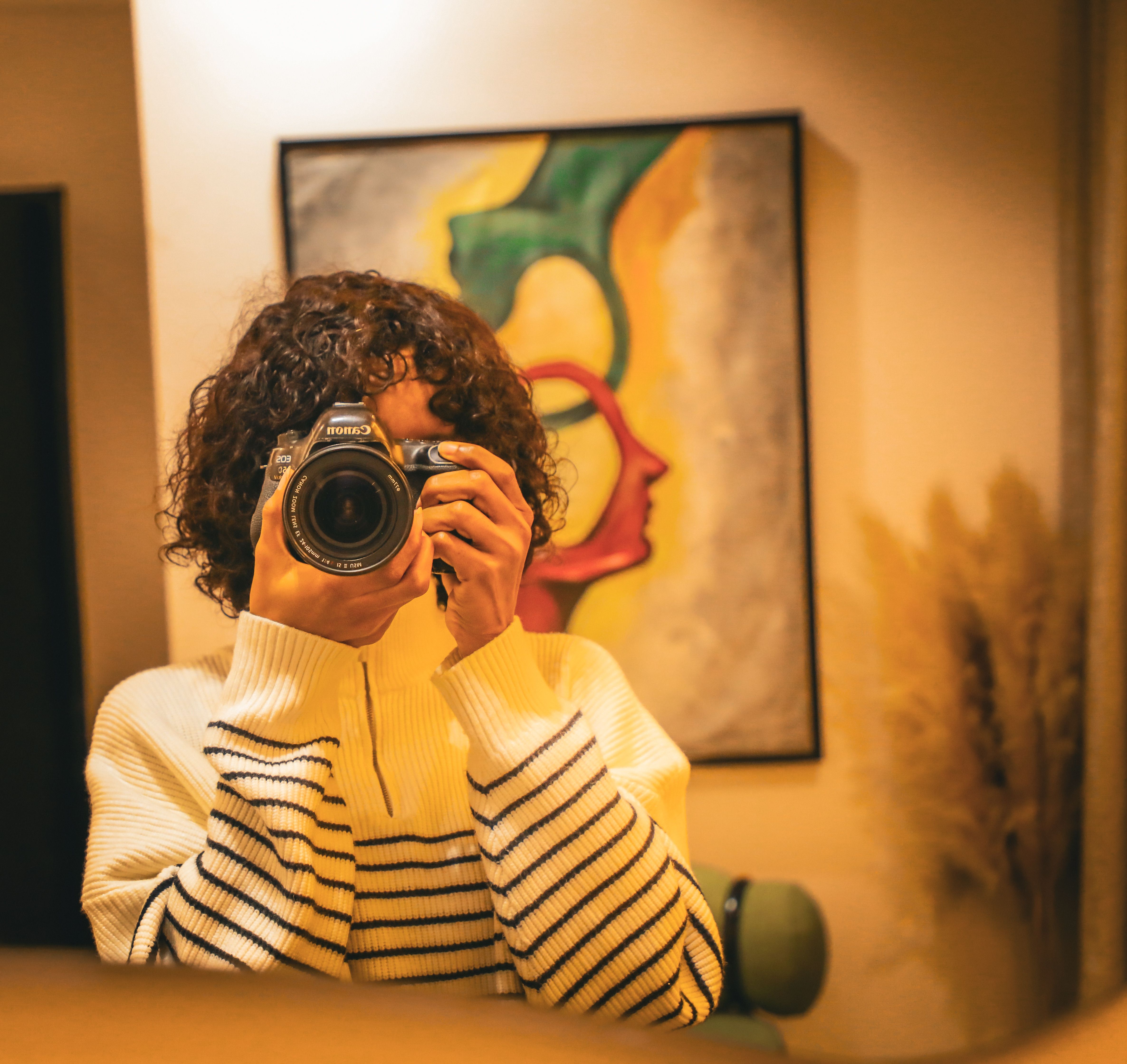 A woman photographs herself in a cozy indoor setting with vibrant art in the background.
