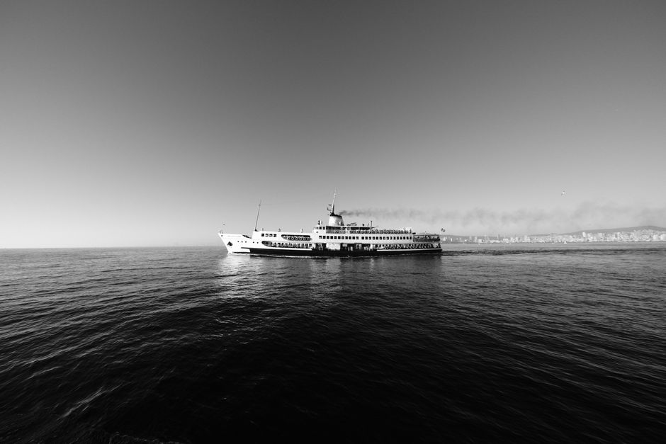 Black and white image of a ferry crossing the open sea with a cityscape in the distance.