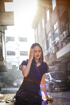 An elegant woman in a black dress poses in a sunlit urban alleyway, showcasing modern fashion.