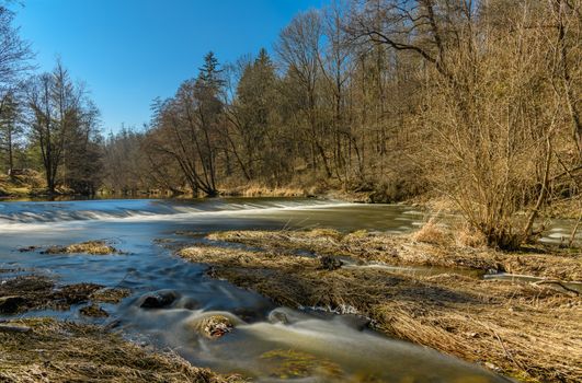 Tranquil river scene with trees and clear blue sky in early spring, showcasing nature's beauty.