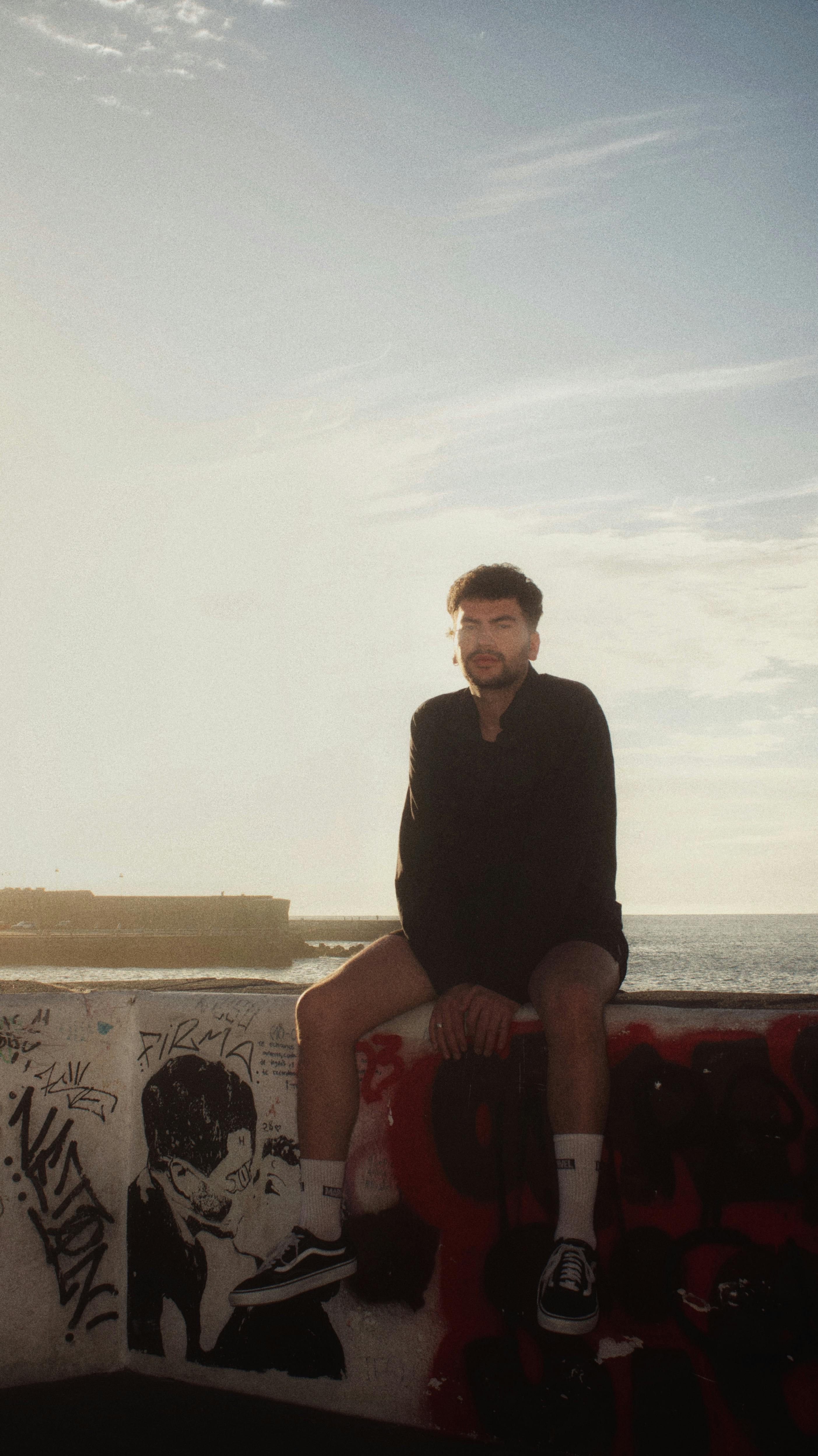 A man sits atop a graffiti-covered wall overlooking the ocean in a relaxed and contemplative pose.