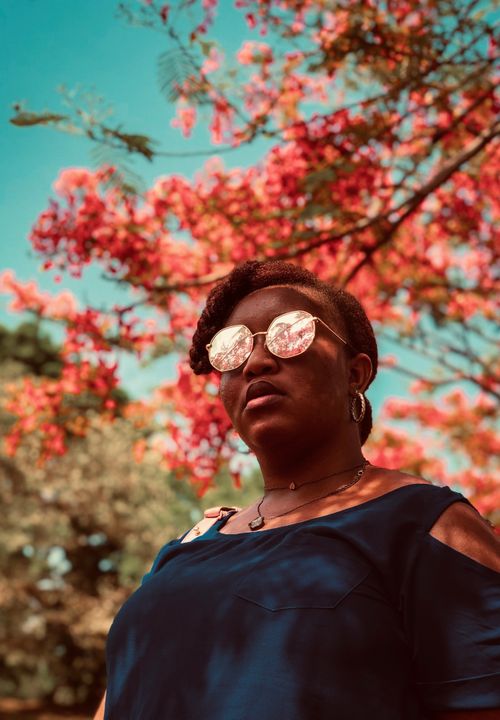 Free A confident woman in sunglasses stands under a tree with bright, colorful blossoms. Stock Photo