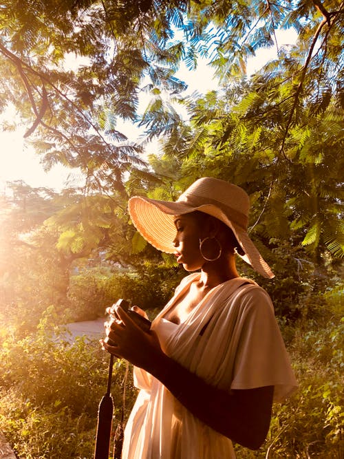 Side view of African American woman in white dress and straw hat standing among branches of green tree and looking on photo camera