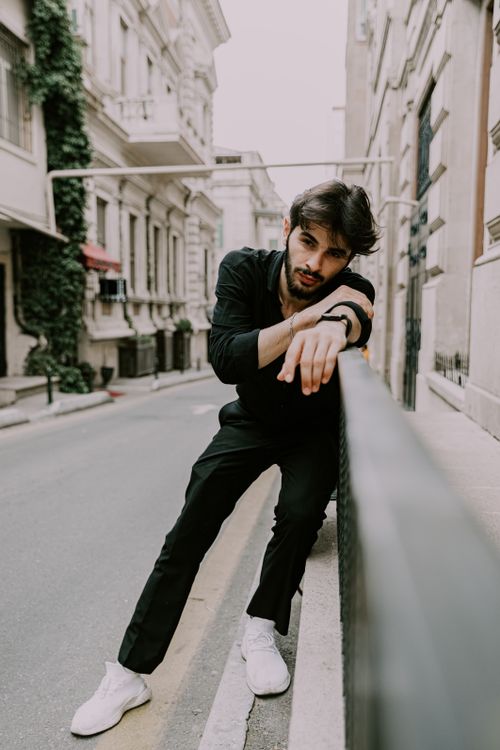Free Fashionable man wearing black outfit and white sneakers leaning on a fence in a Baku street. Stock Photo