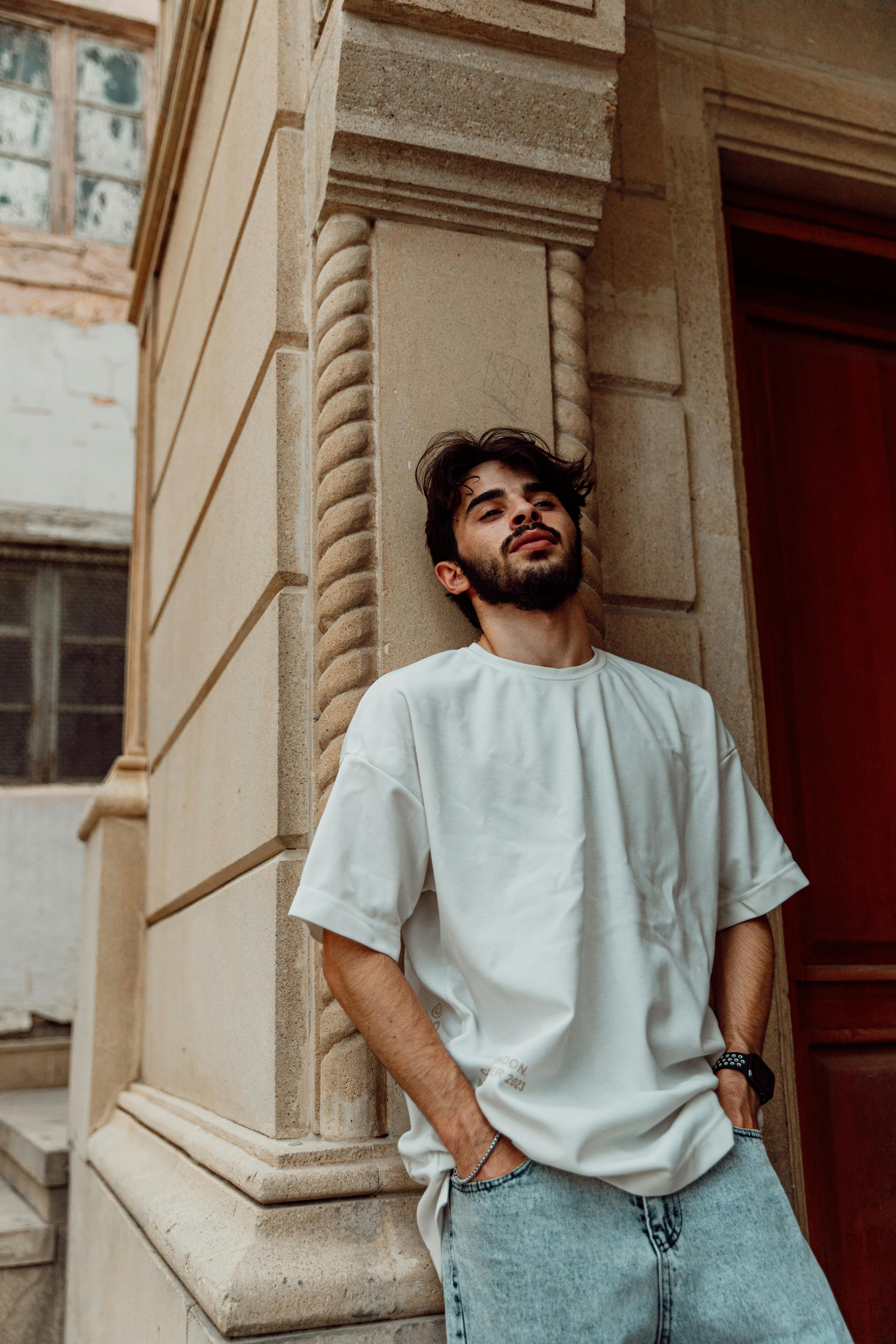 a man leaning against a wall wearing a white t shirt