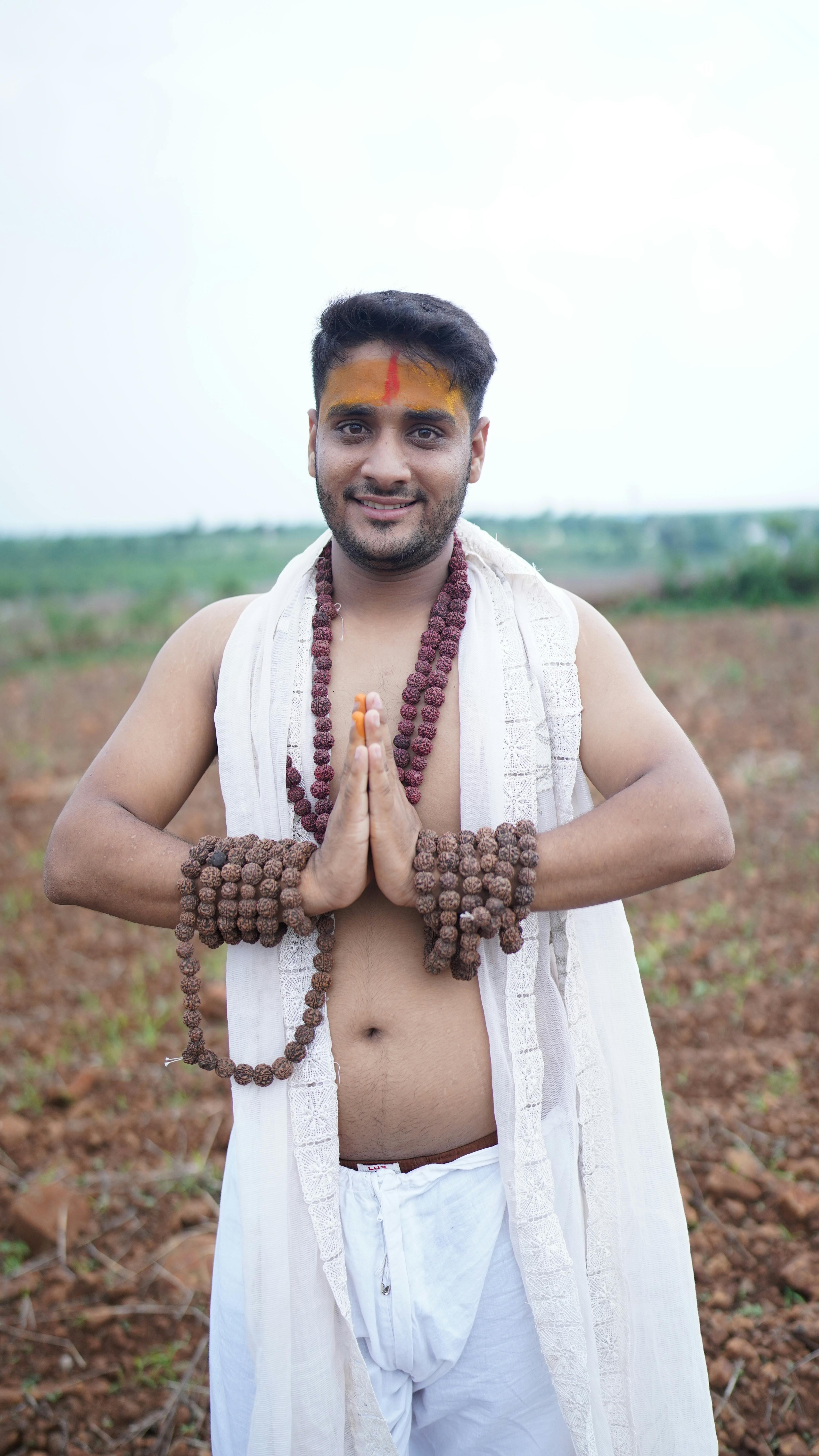 Young man in traditional attire, smiling with namaste gesture outdoors.