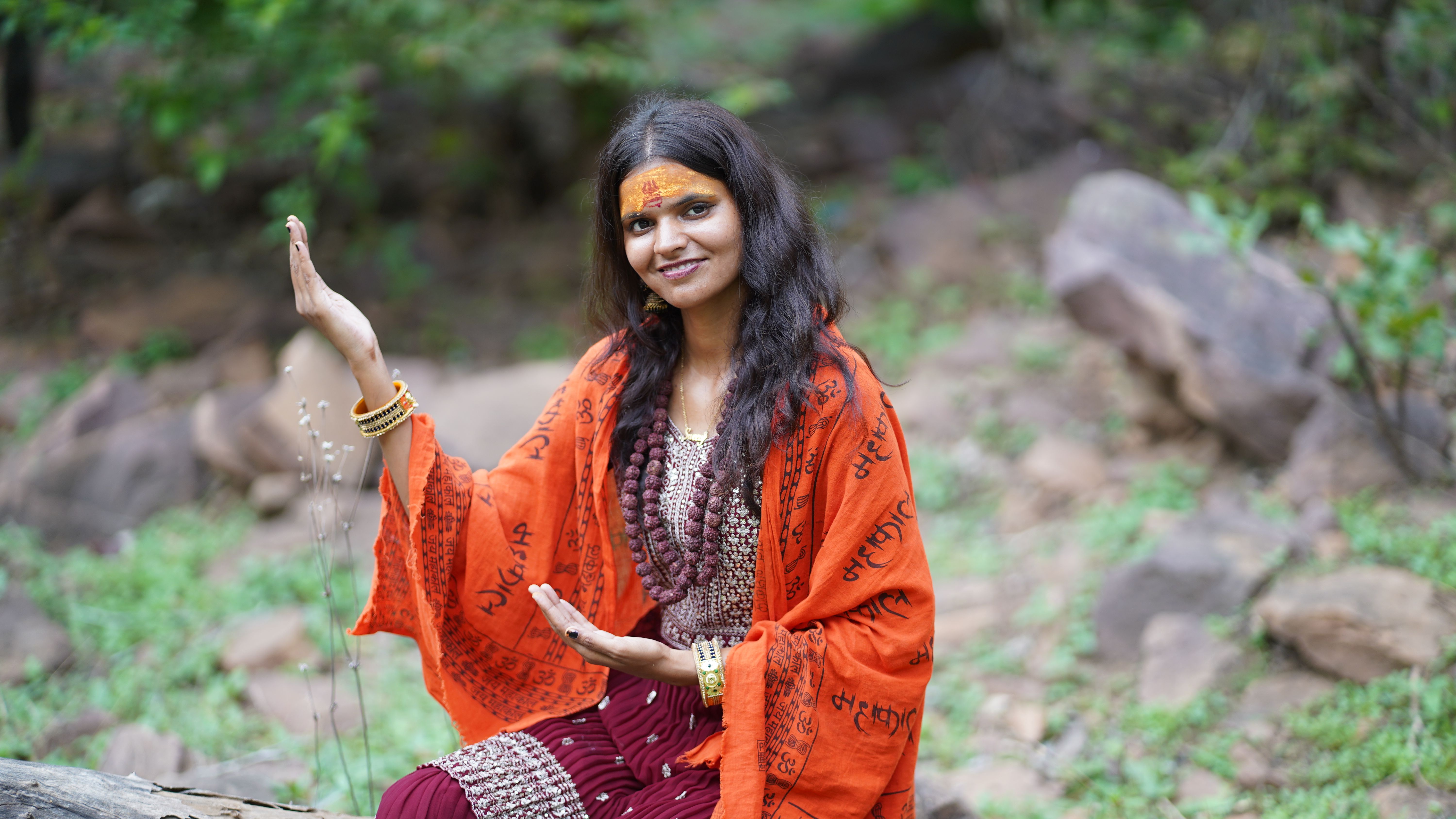 A woman in traditional attire smiling, seated outdoors with a natural backdrop.