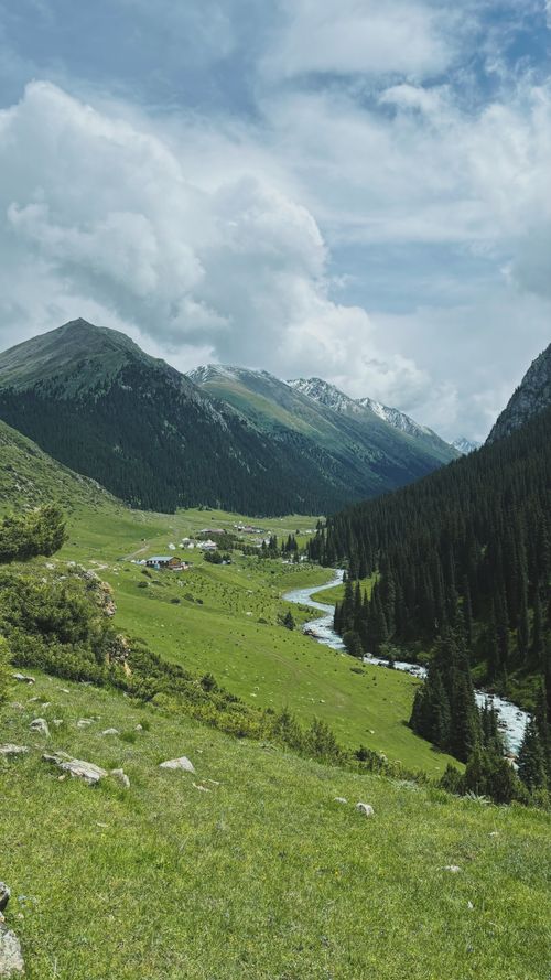 Free Picturesque mountain valley with lush meadows, flowing river, and snow-capped peaks under a cloudy sky. Stock Photo