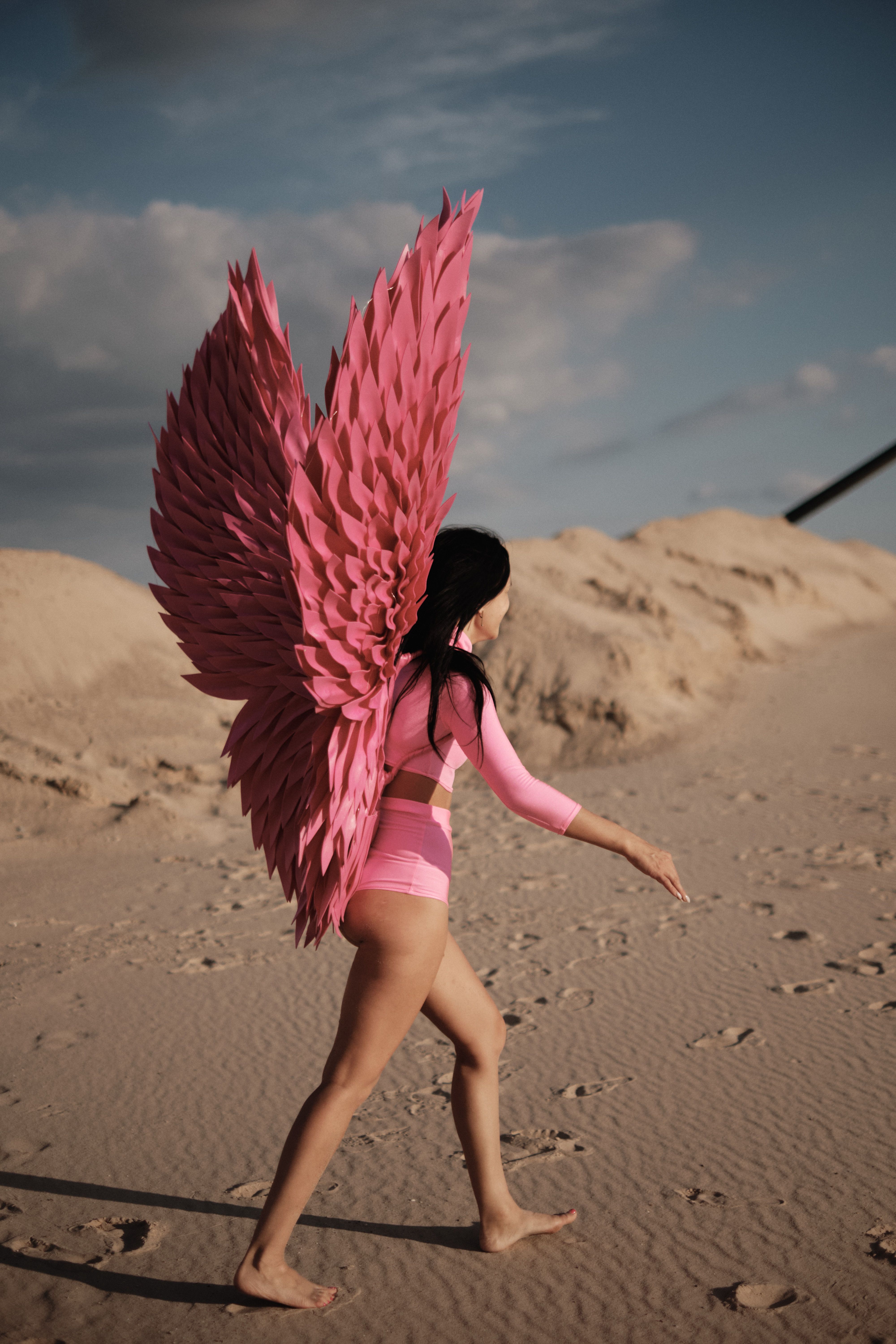 A woman in pink costume with large wings walks on sandy dunes under clear skies.