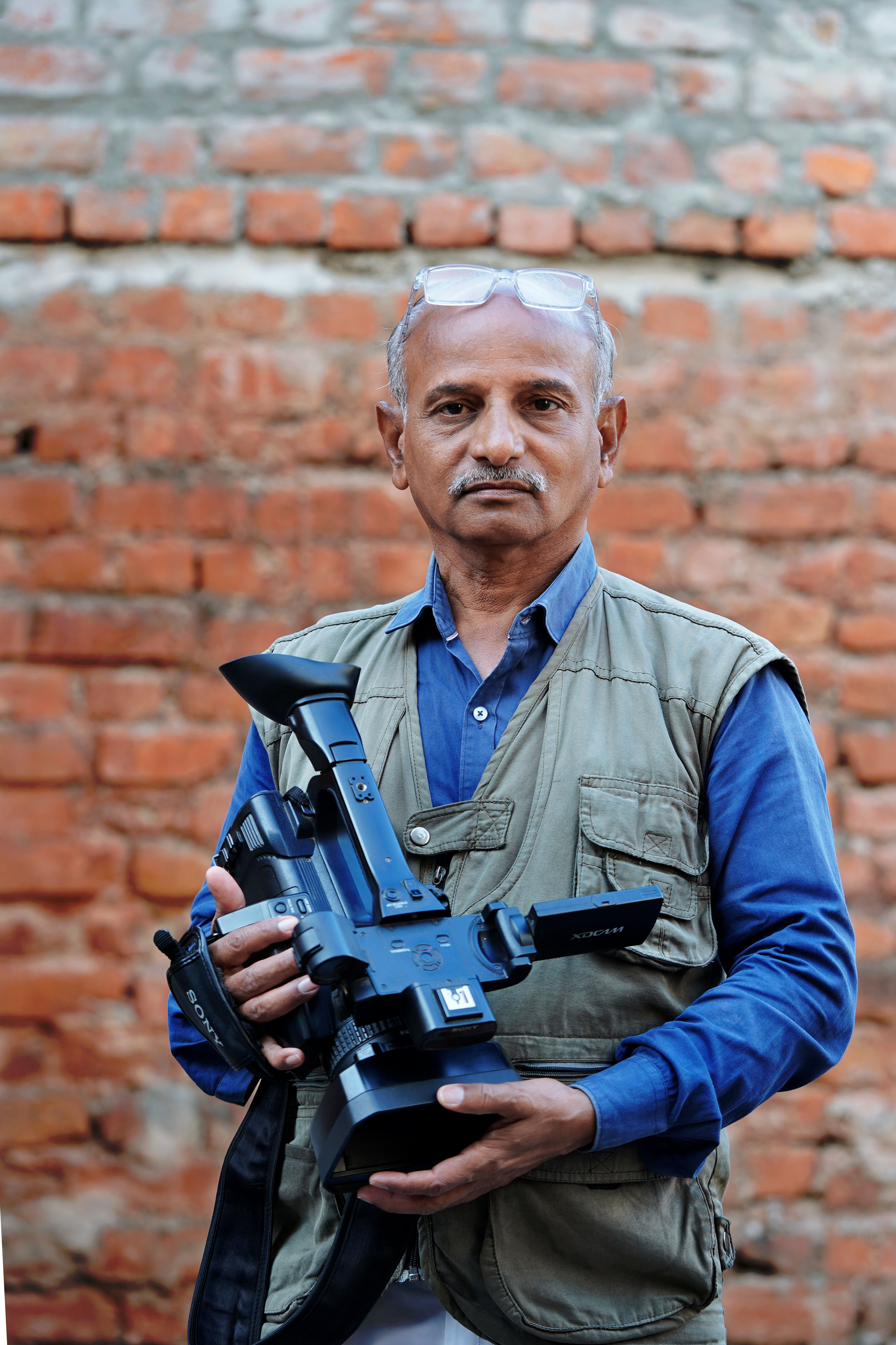 An experienced cameraman holding equipment against a rustic brick wall in an urban outdoor setting.