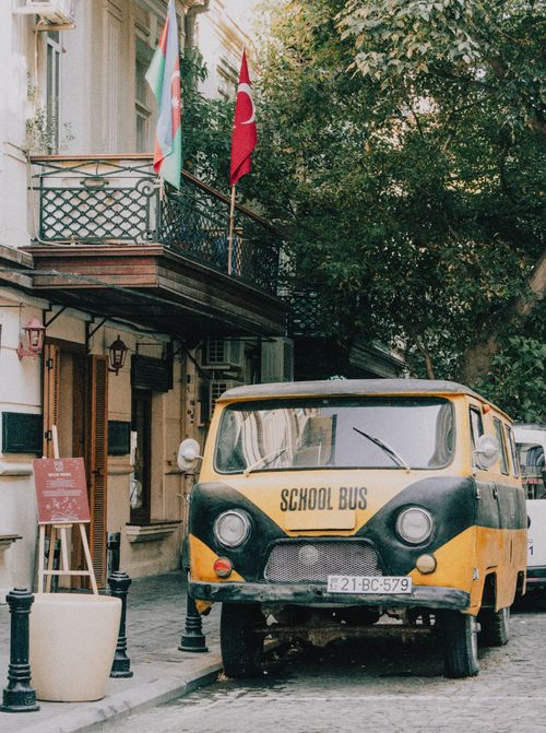 Free A classic yellow school bus parked on a cobblestone street beside a building with national flags. Stock Photo