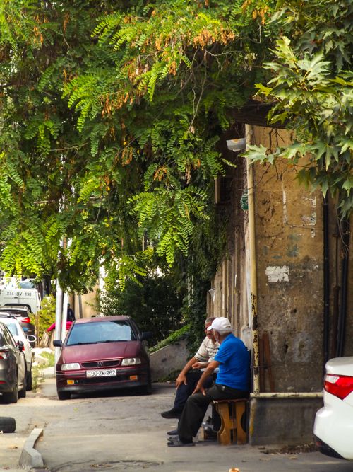 Free Two men sitting on a bench in a shaded street in Baku, Azerbaijan, with cars parked and foliage. Stock Photo