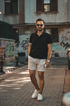 Stylish young man in sunglasses and casual attire strolling on a graffiti-lined urban street.