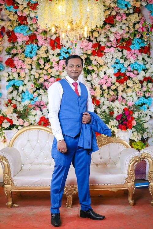 Free A handsome groom in blue wedding attire poses in front of a vibrant floral backdrop. Stock Photo