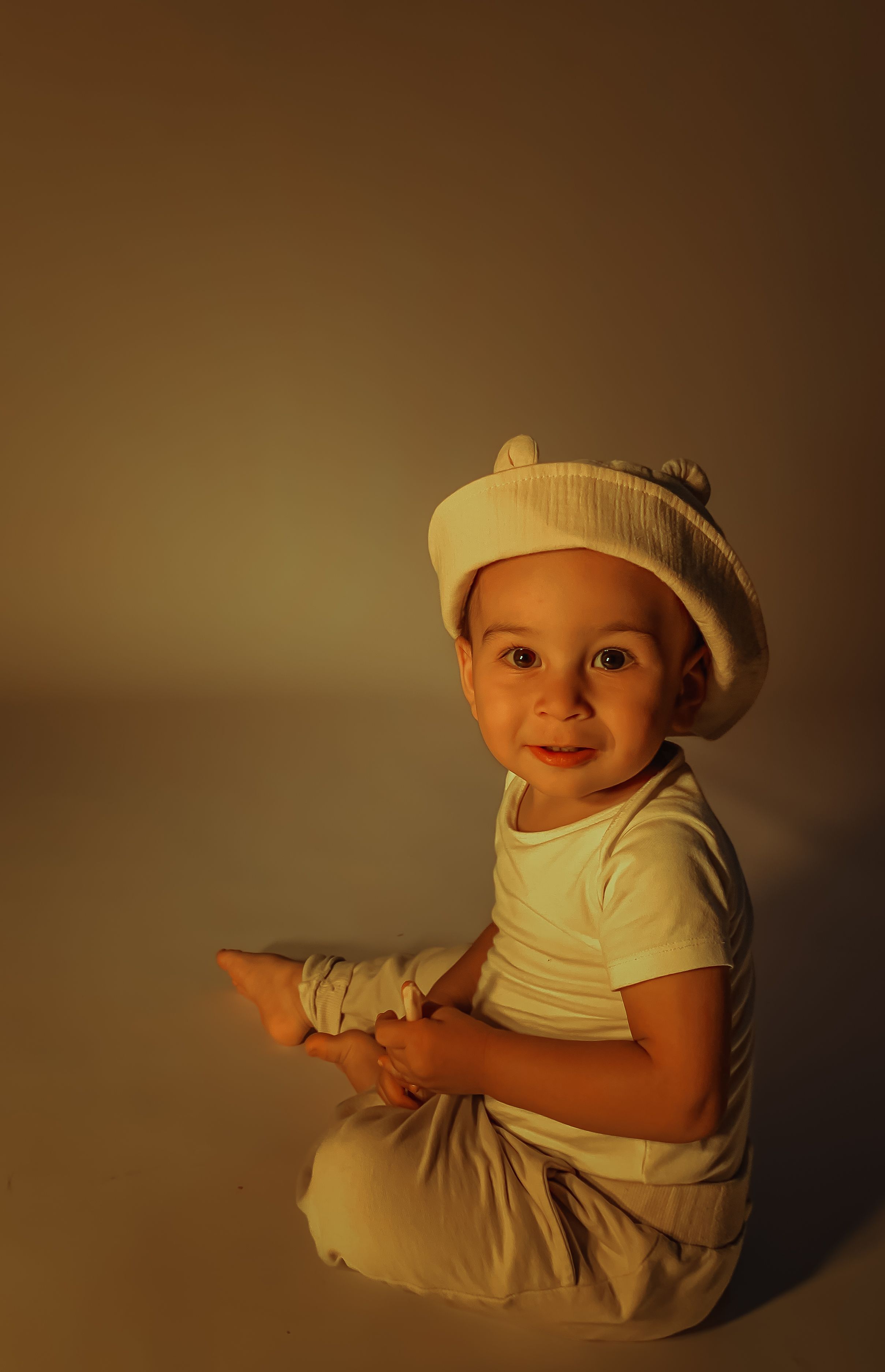 Adorable child sitting in a studio, wearing a cap with warm lighting. Captures innocence and joy.