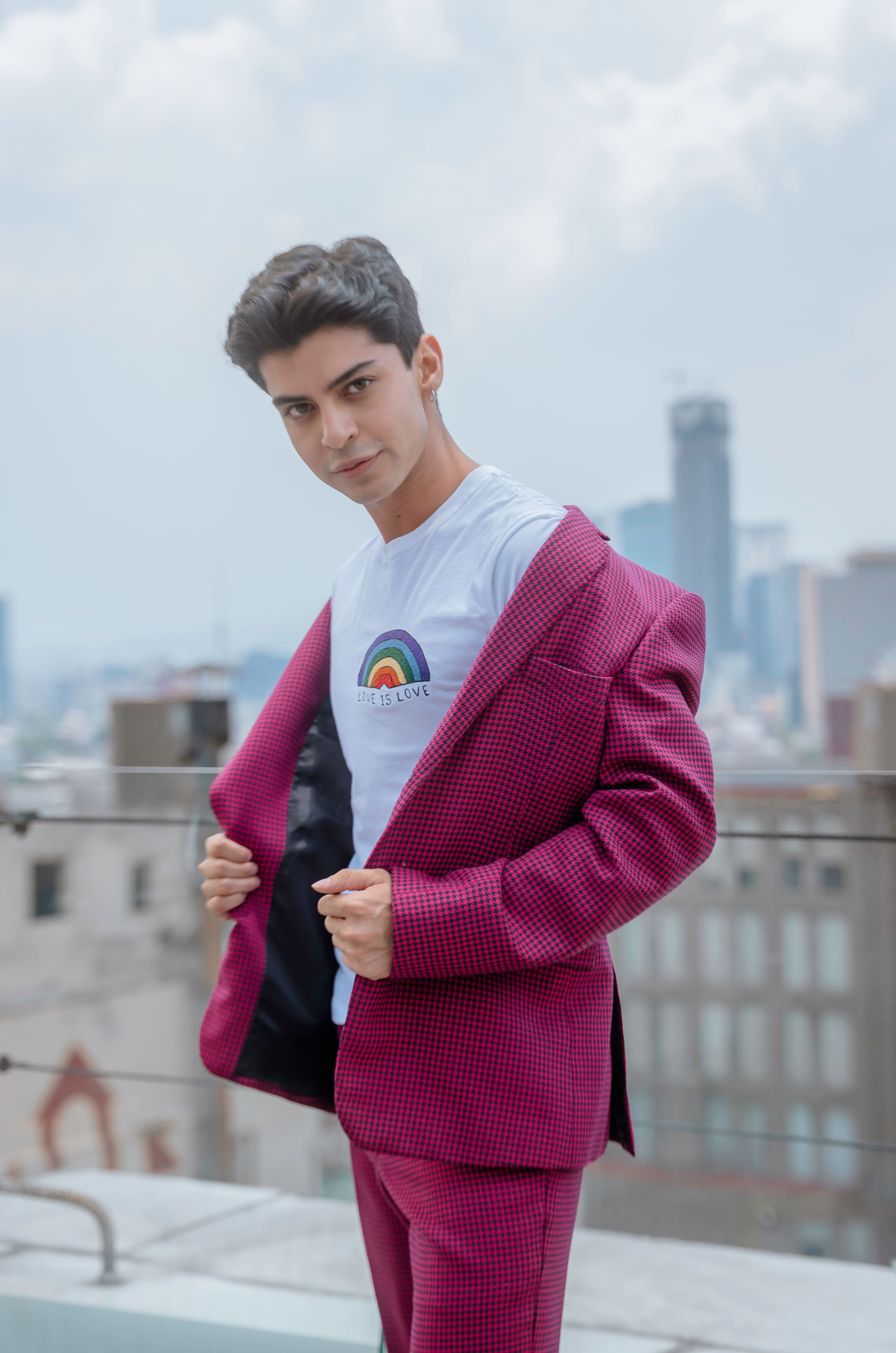 Confident young man poses in a pink suit on a rooftop with a view of Mexico City's skyline.