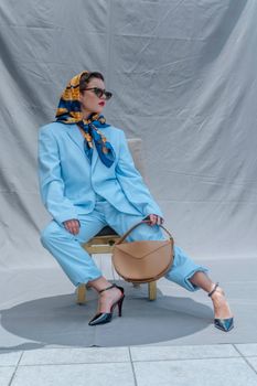 Fashionable woman in a blue suit and scarf, holding a bag and sitting stylishly.