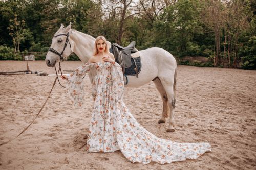 Free A woman in a floral gown poses gracefully with a white horse in a sandy outdoor setting. Stock Photo