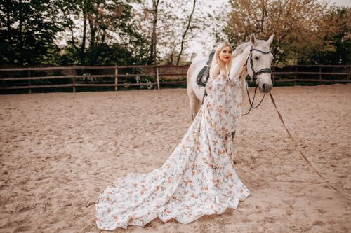 Free Stunning portrait of a woman in a floral dress standing beside a white horse on a sandy outdoor arena. Stock Photo