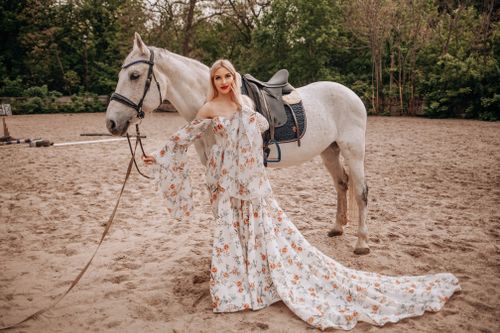 Free A stylish woman poses with a horse in an elegant floral dress on a sandy path. Stock Photo
