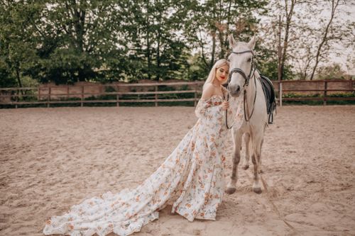 Free A beautiful woman in a floral dress standing beside a horse in an outdoor arena. Stock Photo