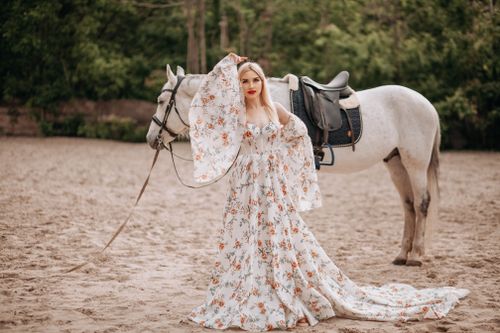 Free A woman in a floral dress poses gracefully with a white horse in a serene outdoor setting. Stock Photo