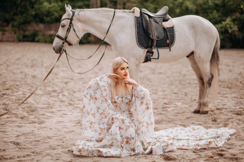Free Elegant woman in floral dress sitting by a white horse with a saddle on sandy ground. Stock Photo