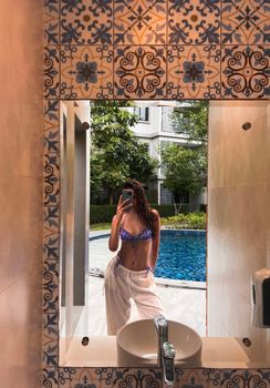 Woman in bikini and pants takes a selfie in a bathroom mirror with pool view.