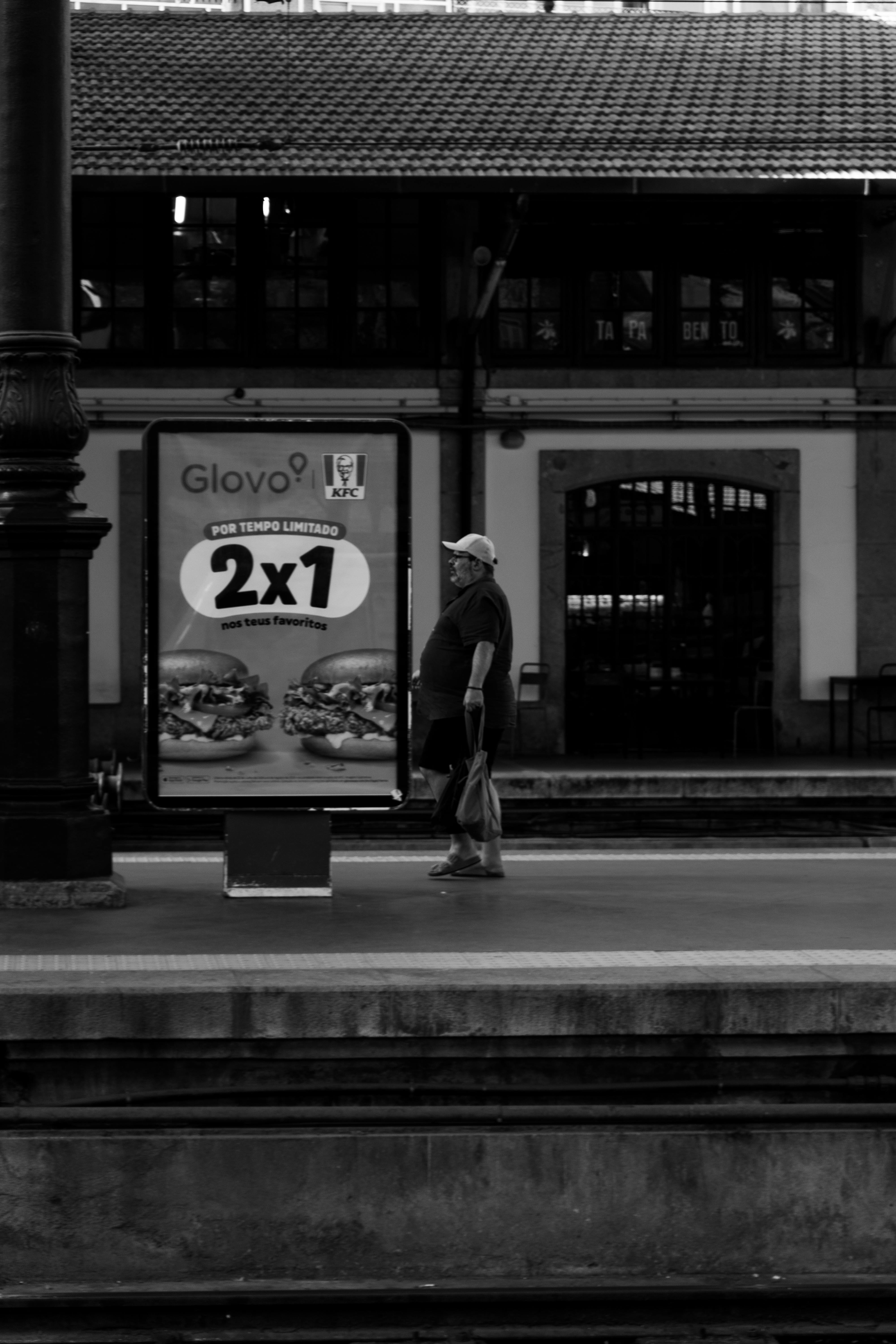 Black and white photo of a man waiting by an ad at a Porto train station.
