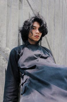 A young man in a stylish apron poses against a concrete wall. Urban fashion portrait.