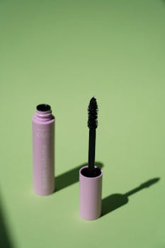 Open mascara tube with brush resting on vibrant green background, showcasing cosmetic product.