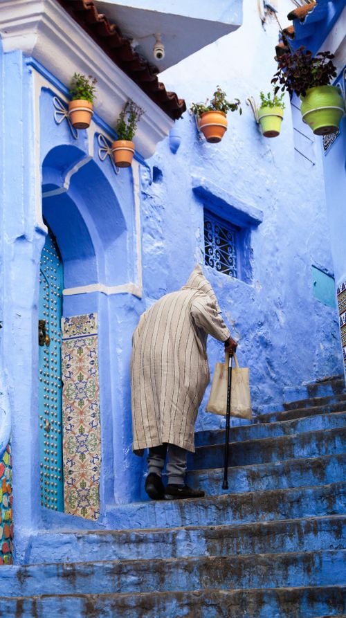 Free An elderly man climbs the vibrant blue stairs of Chefchaouen, Morocco, showcasing local architecture. Stock Photo