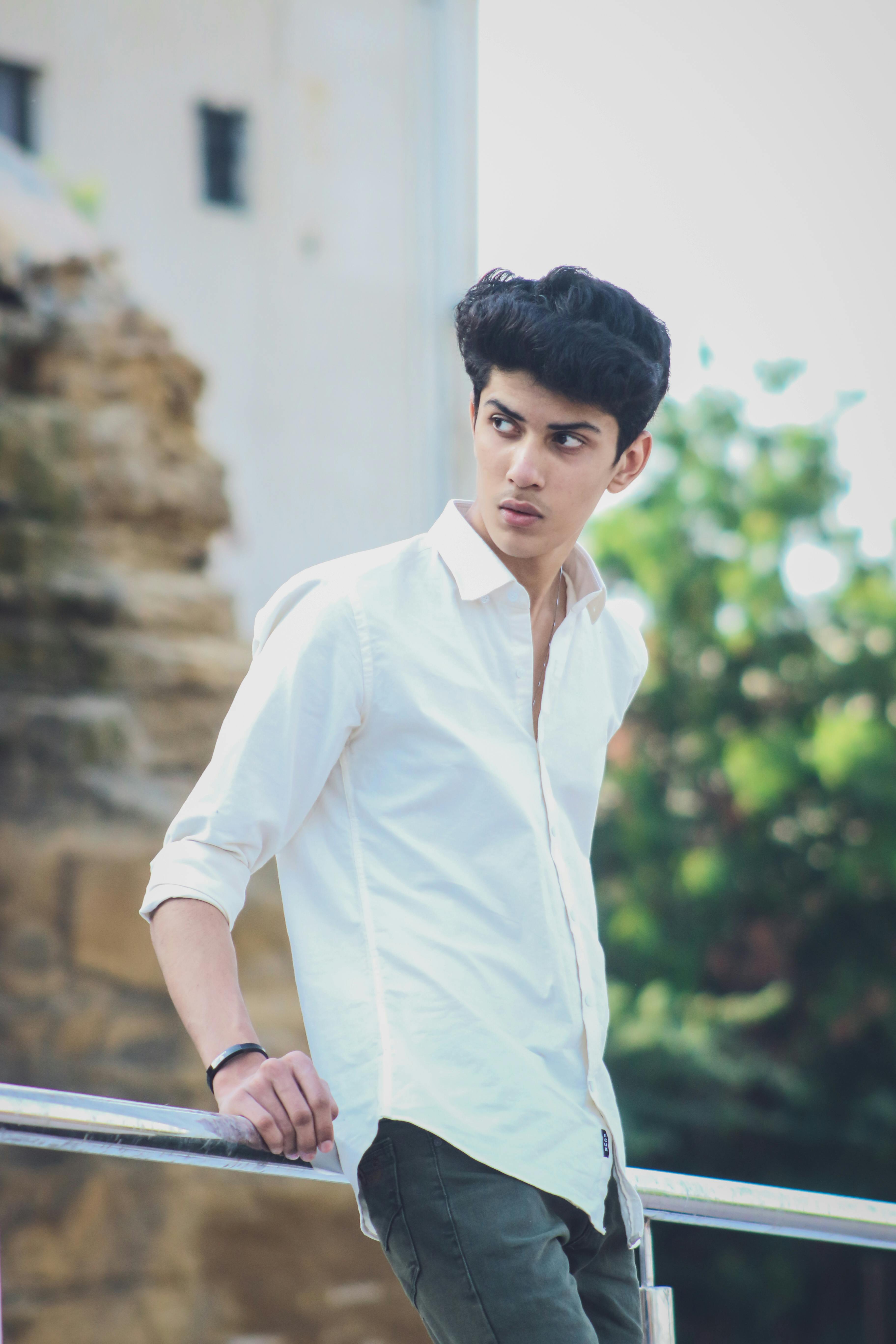 A young man in a white shirt and casual attire leans outdoors, looking thoughtful.