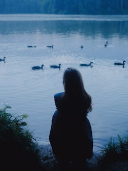 A woman sits by a tranquil lakeside with ducks swimming at sunrise in Belarus.