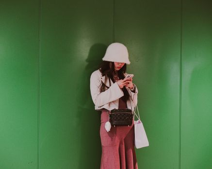 Fashionable woman in a chic hat using a smartphone against a green background in Tokyo.