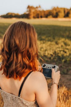 A woman operates a drone controller during a warm, golden sunset in a rural field, capturing the serene summer landscape.
