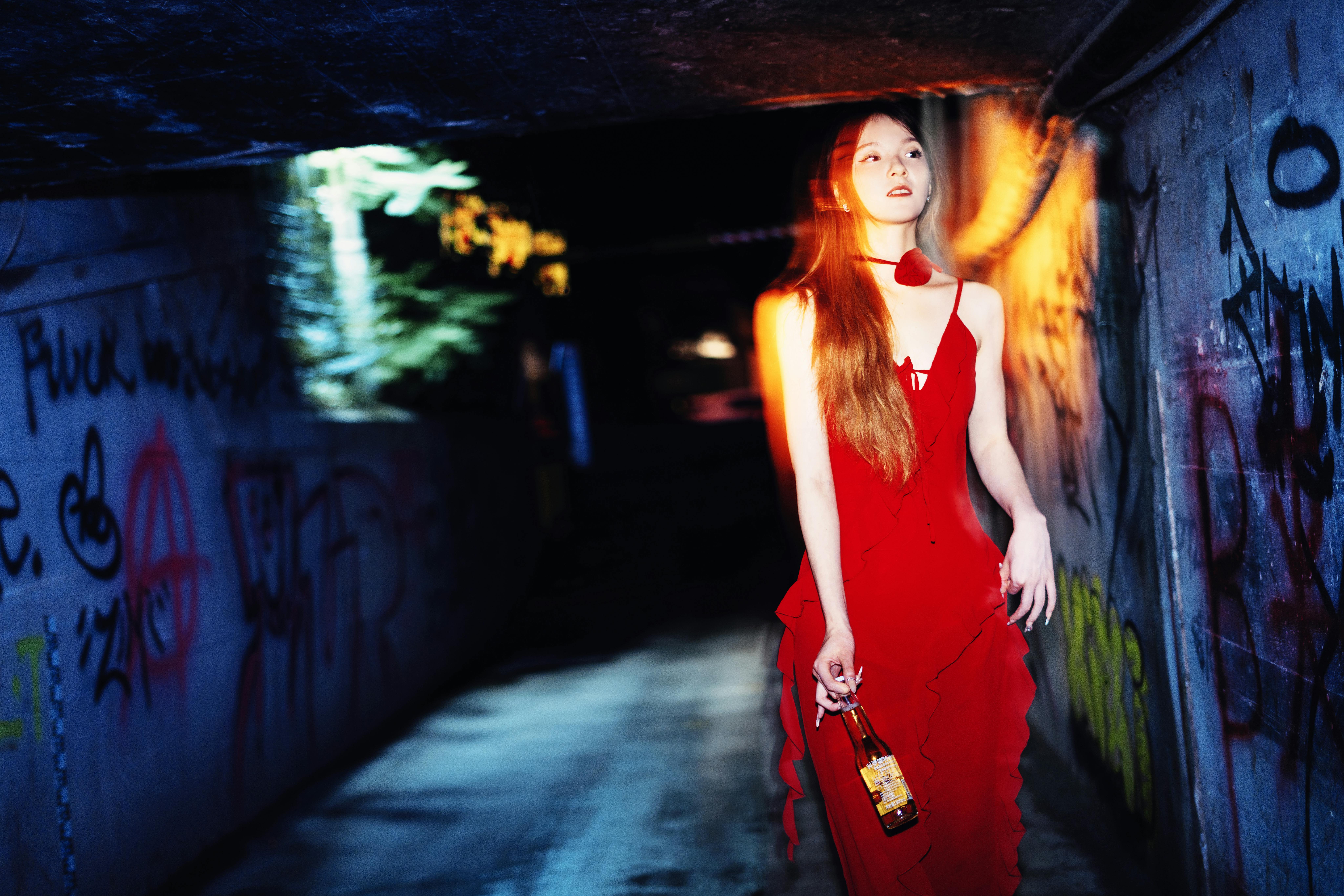 A young woman in a red dress walks through a graffiti-covered alleyway at night.