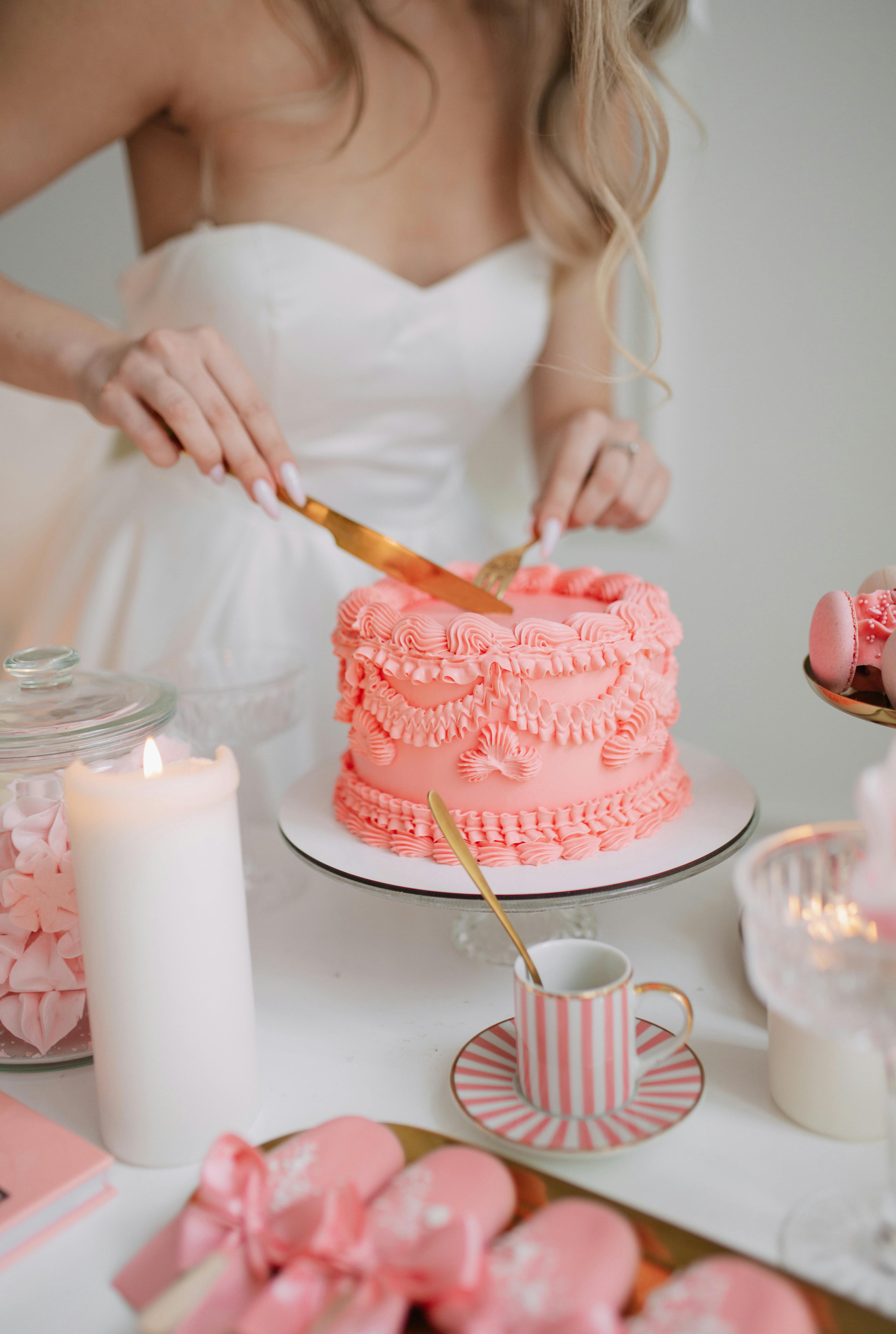 A woman in a white dress decorates a pink cake beside candles and macarons in an elegant setting.