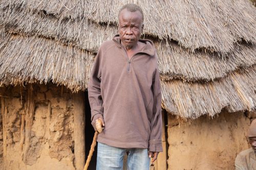 Free An elderly man stands in front of a rustic mud hut with a thatched roof, showcasing rural life. Stock Photo
