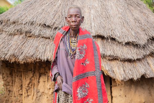 Free Portrait of an elderly indigenous woman standing in front of a rural thatched hut showcasing traditional attire. Stock Photo